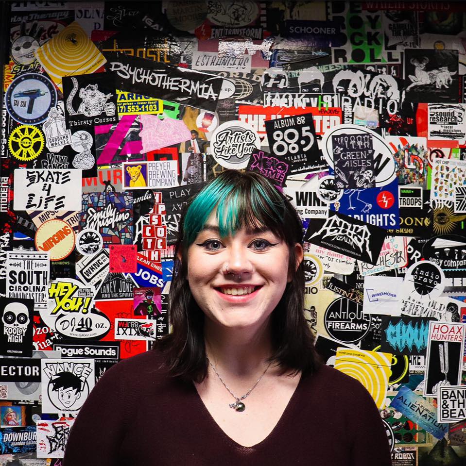 Person with green hair standing in front of a heavily stickered door