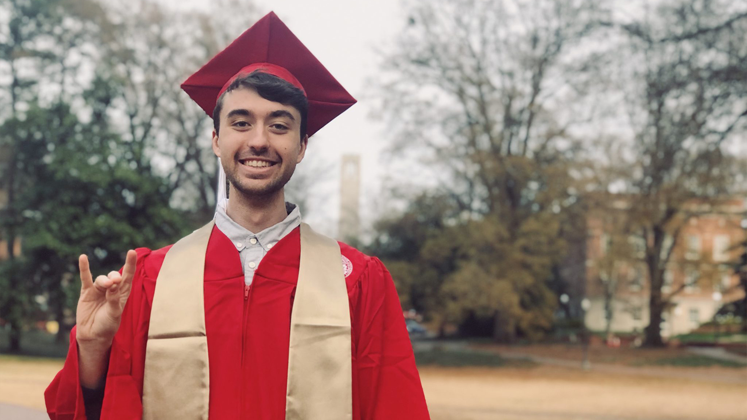 Colton Botta in red graduation robes making a wolfie sign with the bell tower in the background