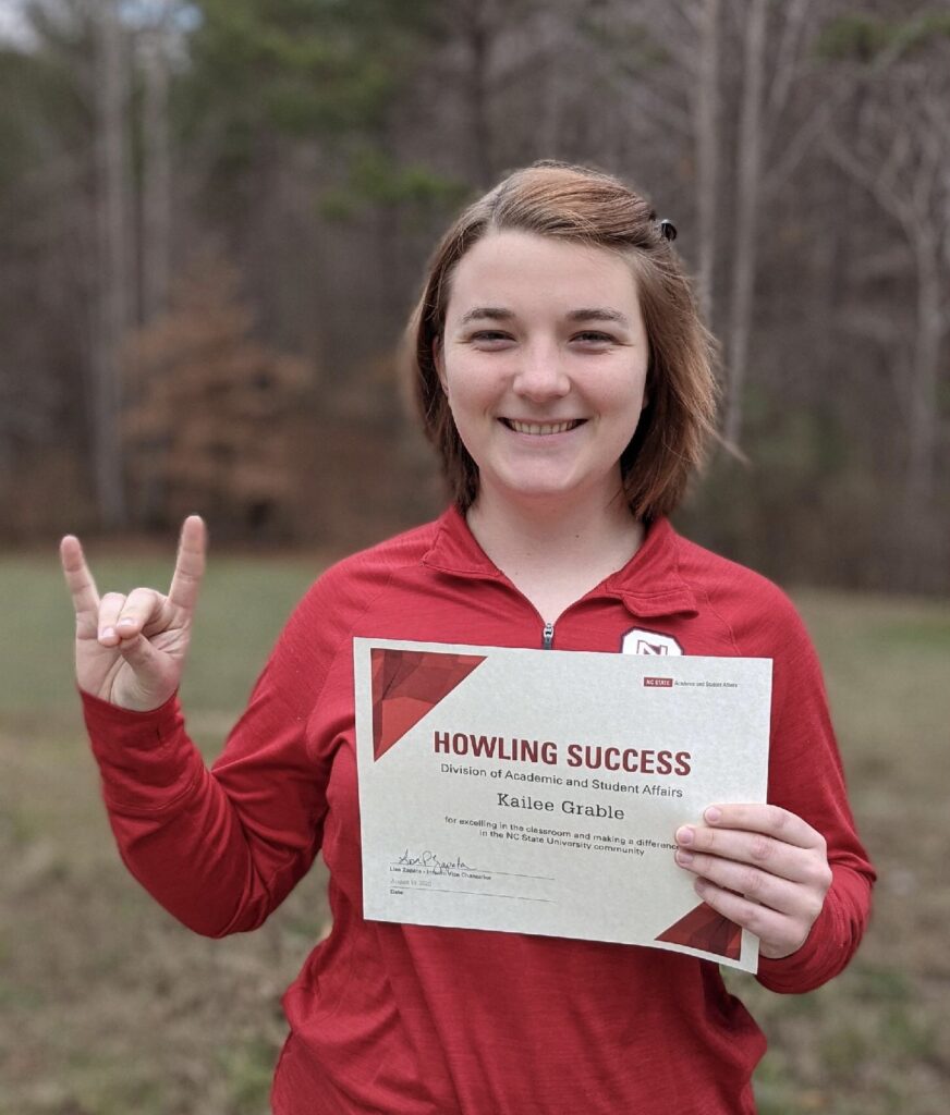 Kailee Grable, wearing a red jacket, holding a certificate and making a Wolfie sign with her hand