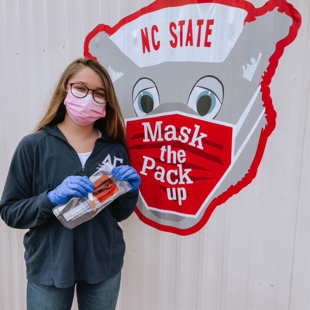 A masked, female student holds a test kit in front of a sign that reads "Mask the Pack Up"