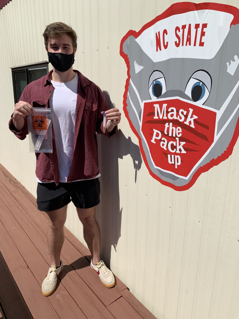 A male, masked student holds a COVID-19 test kit next to a sign that reads "Mask the Pack Up."