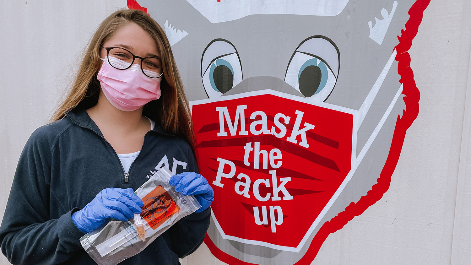 A female student holds a COVID-19 test bag in front of a Mr. Wuf head sign that reads "Mask the Pack Up"