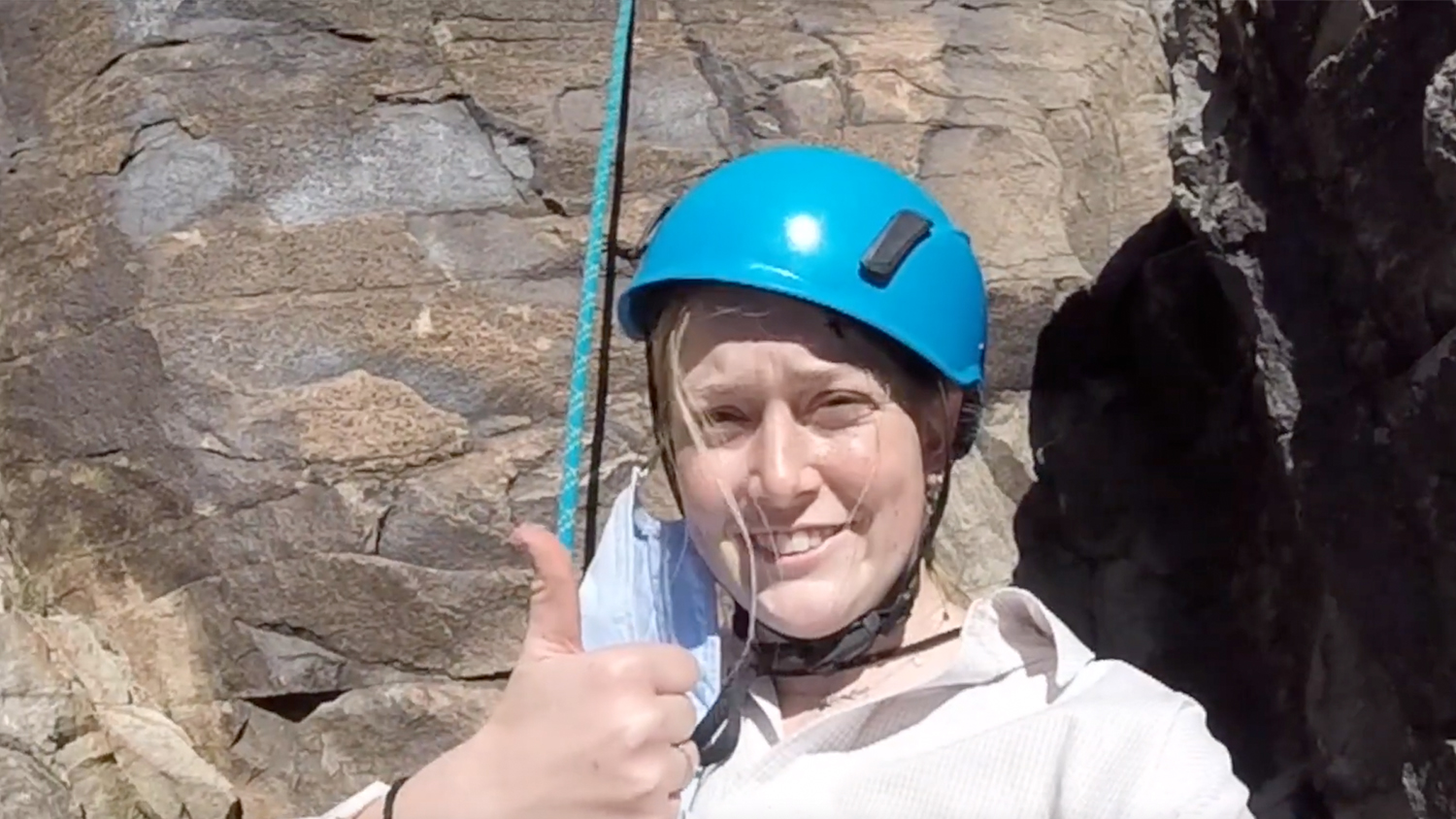 Massey Stichter wearing a teal helmet and making a thumbs up sign while rock climbing
