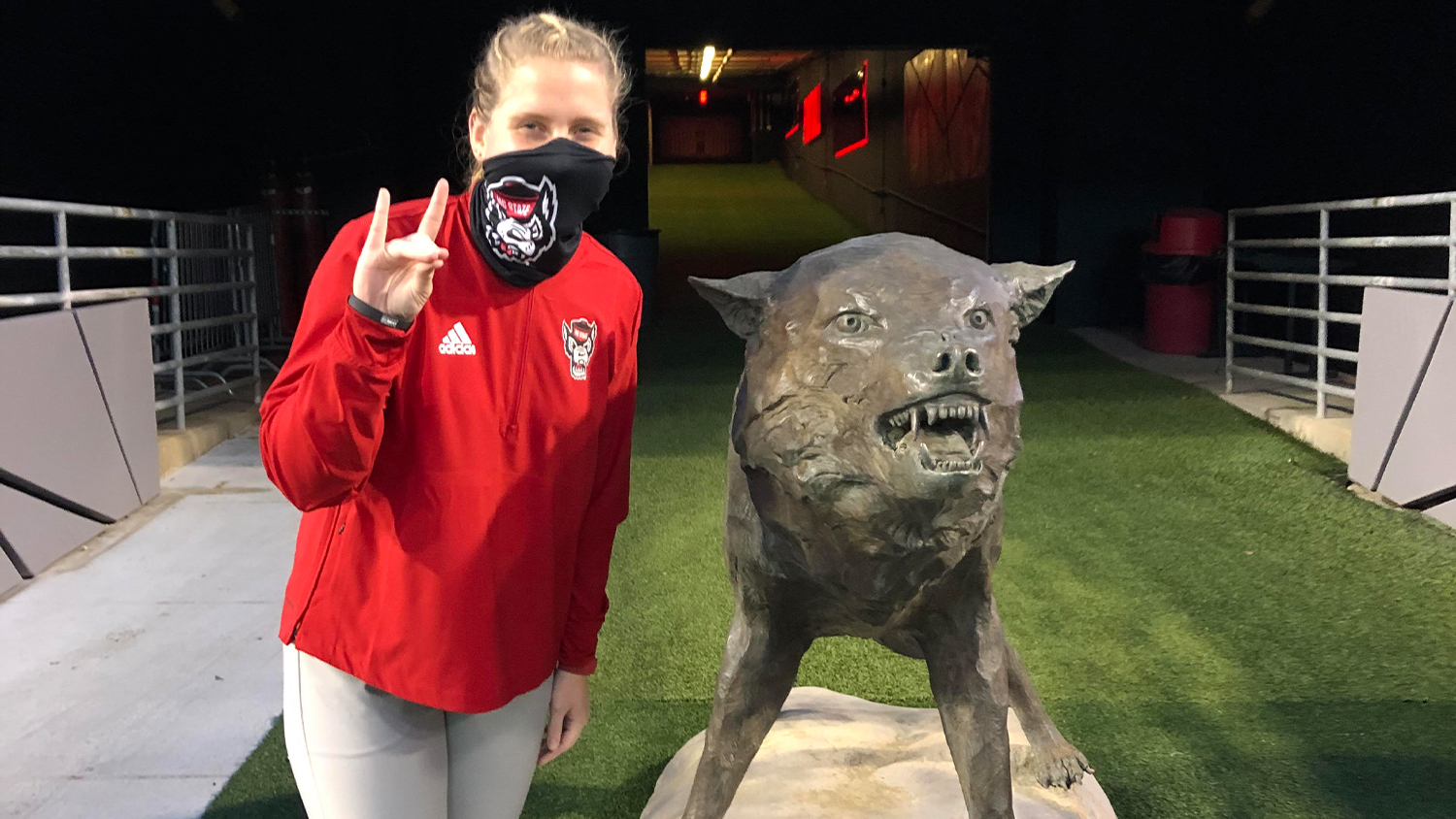 Crista Bain wearing a red jacket and a black NC State face mask makes a wolfie sign next to the wolf statue in Carter Finley Stadium
