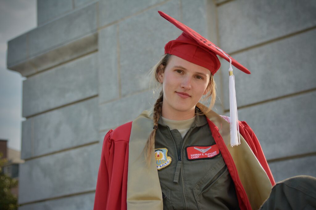 Hannah Fletcher in red graduation robes and red cap, with Air Force flight suit underneath, standing at the base of the bell tower
