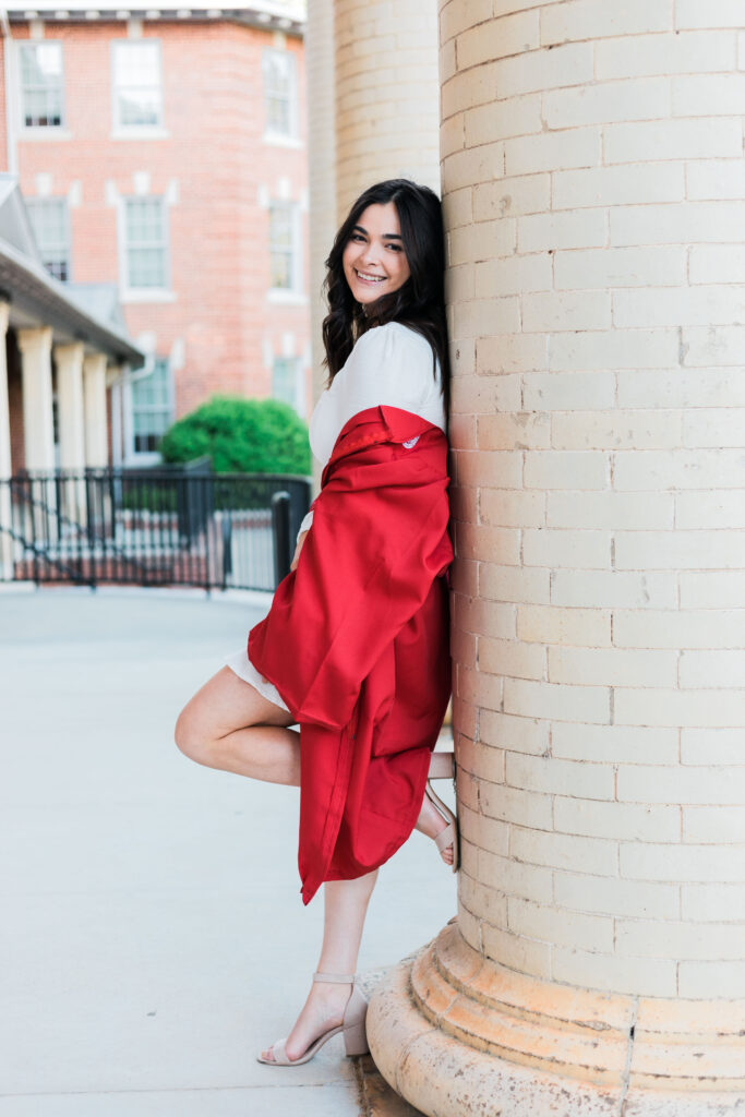 Gillian Torr, wearing a white dress draped in red graduation robes, leaning against the column of a building on campus