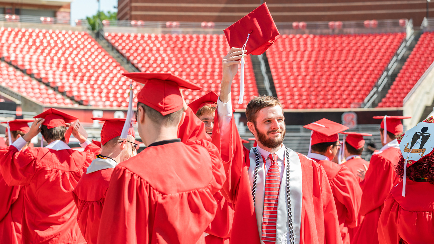 Among several graduates in red robes and hats, one male graduate holds his hat up in the air and smiles
