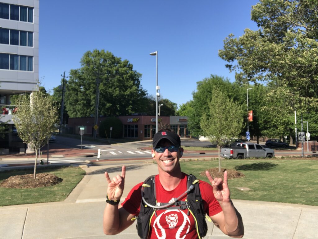 Travis Newton, wearing a red T-shirt, black hat and sunglasses, makes a wolfie sign on a sidewalk on campus