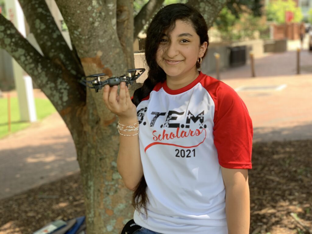 A student in a shirt that reads "STEM Scholars 2021" smiles and holds up their drone 