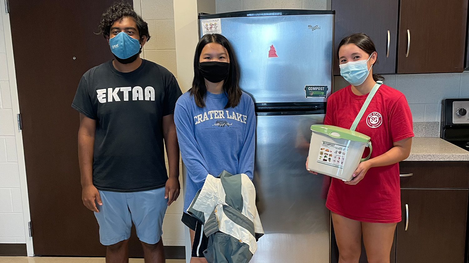 Three students stand in an apartment kitchen, with one holding a compost bucket