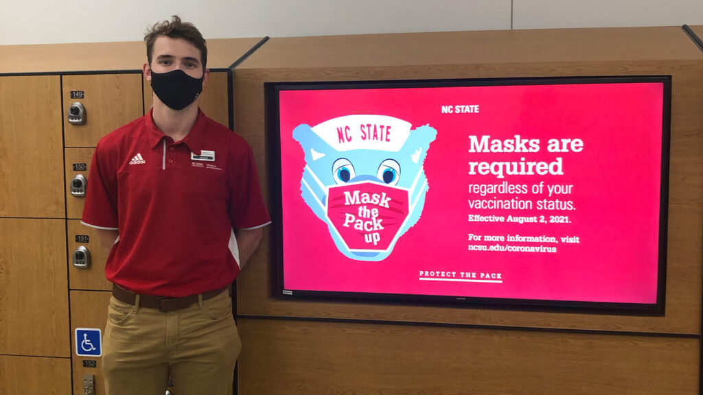 Owen Hanna, stands next to lockers wearing a black face covering and a red polo shirt, standing next to a sign that says "Masks are required regardless of your vaccination status."