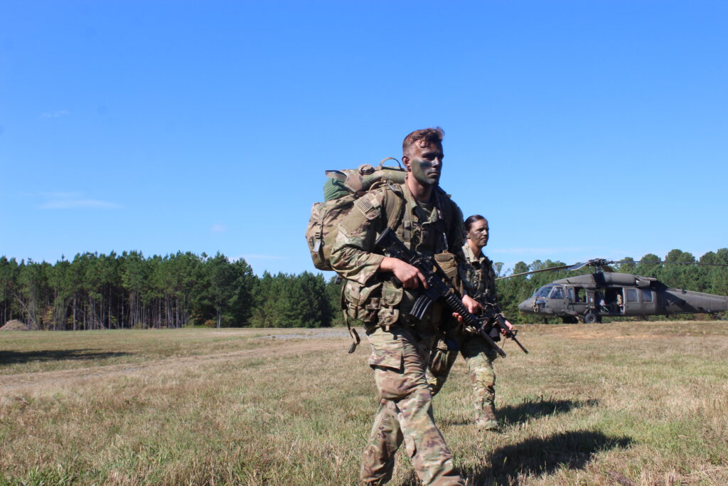 Cadets in full camouflage hold M4 rifles as they walk through a field in front of a helicopter