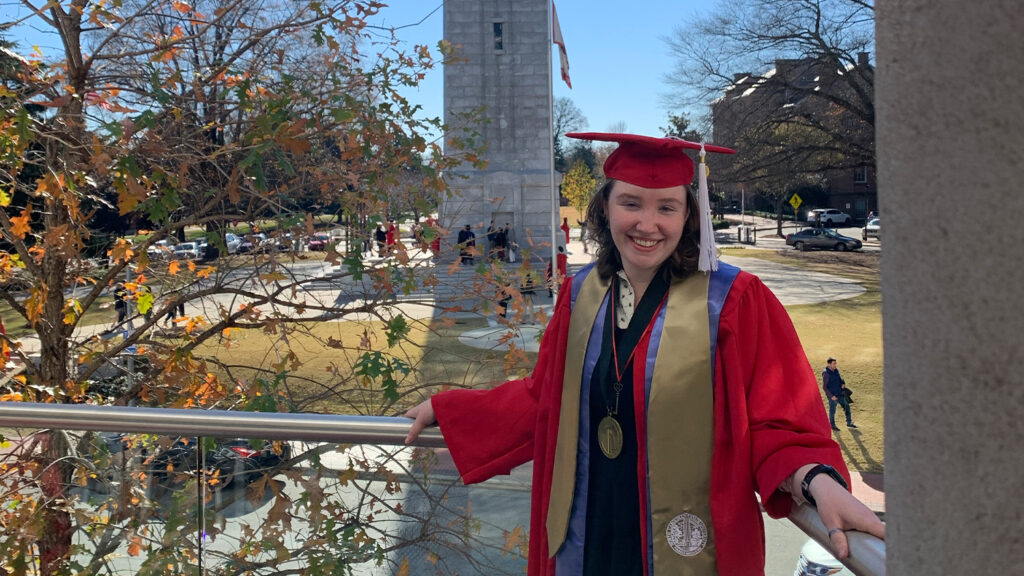 Madison Horgan in red NC State graduation robes, standing on a balcony overlooking the NC State bell tower