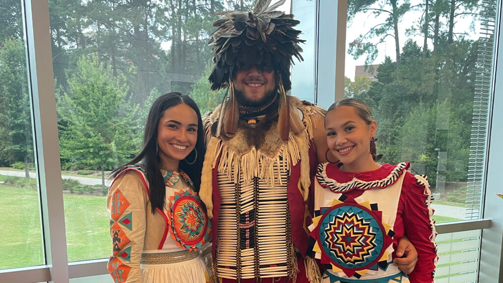 Three students dressed in traditional Native American outfits in front of a large window in Talley Student Union