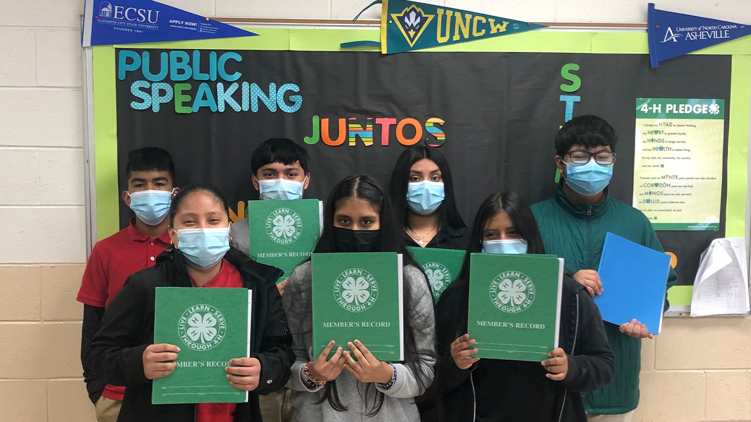 A group of masked students in front of a bulletin board hold up green folders with the 4-H logo on them