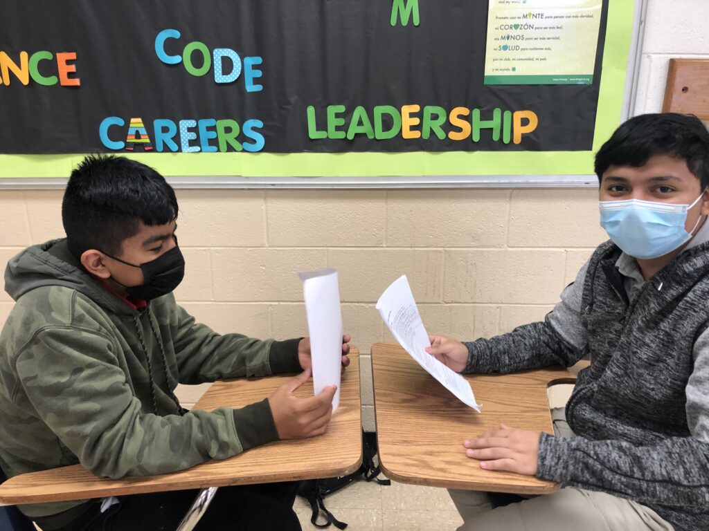Two students sit in their desks facing one another and holding sheets of paper