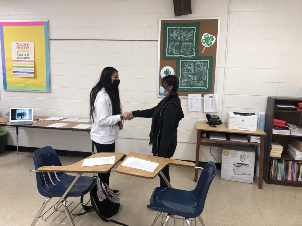 Two students standing next to their desks and shaking hands