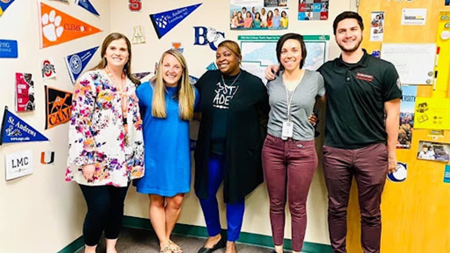 Five members of the Heide Trask High School staff in front of a wall covered in pennants from different colleges and universities