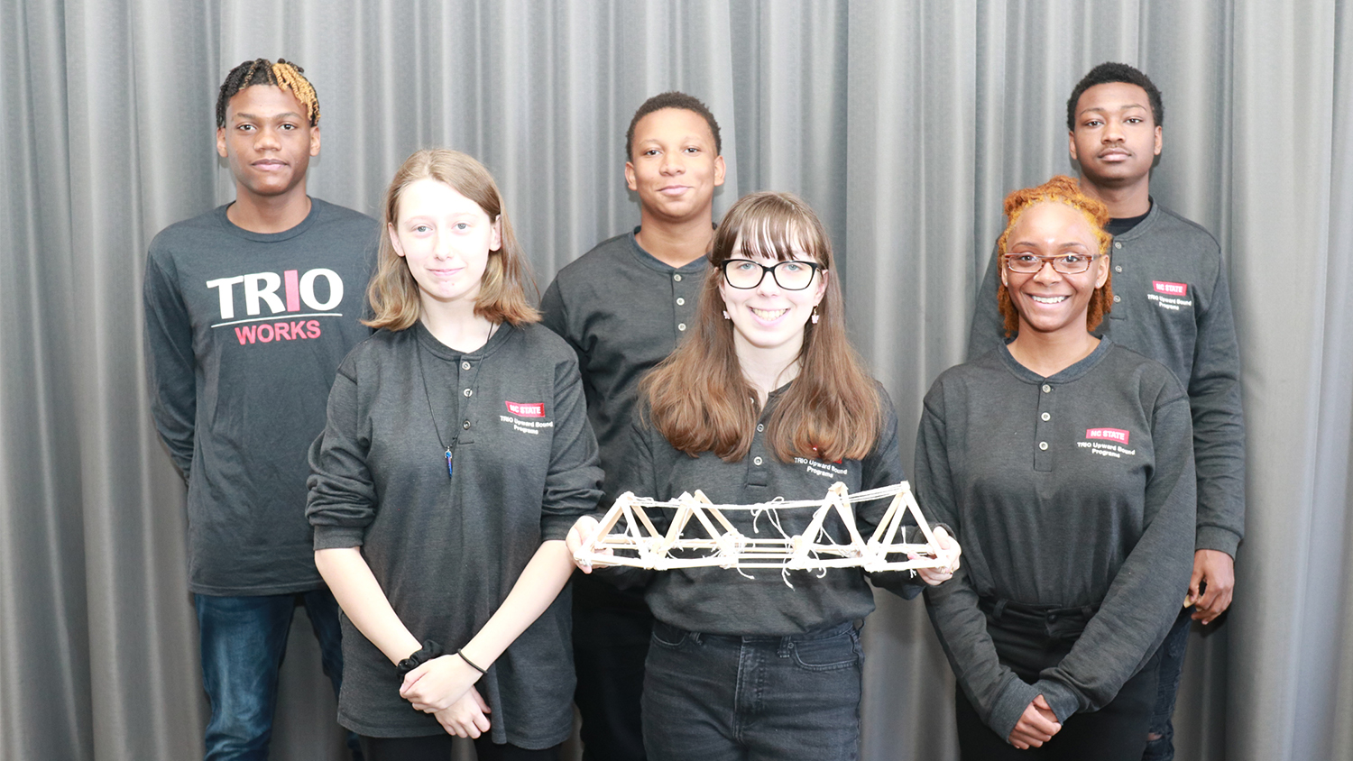 A group of students in grey TRIO T-shirts, with the student in front holding the bridge they built