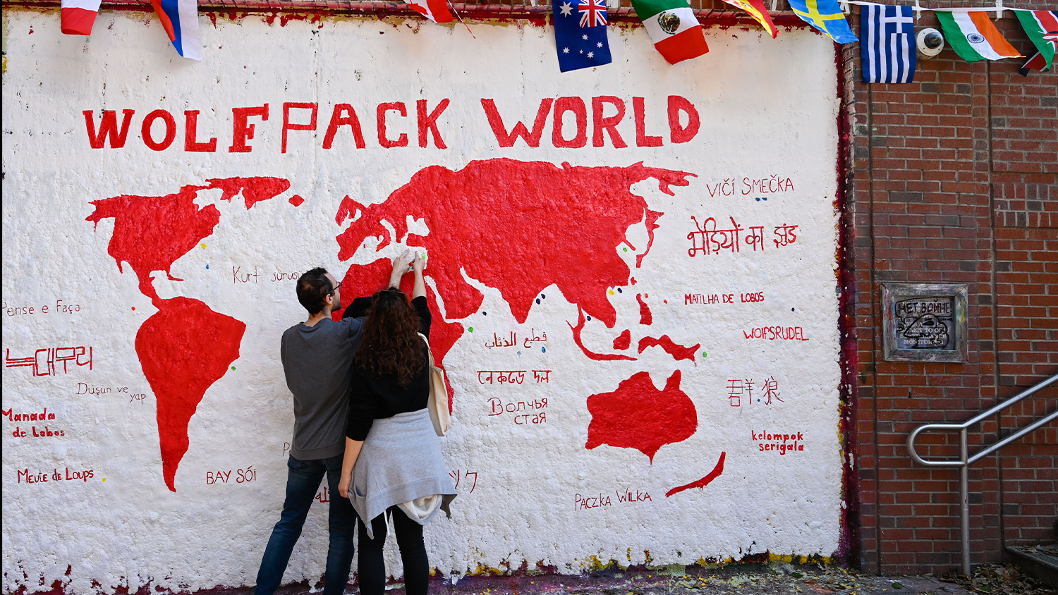 Students add to a mural on the Free Expression Tunnel that shows a map of the world with the text "Wolfpack World"