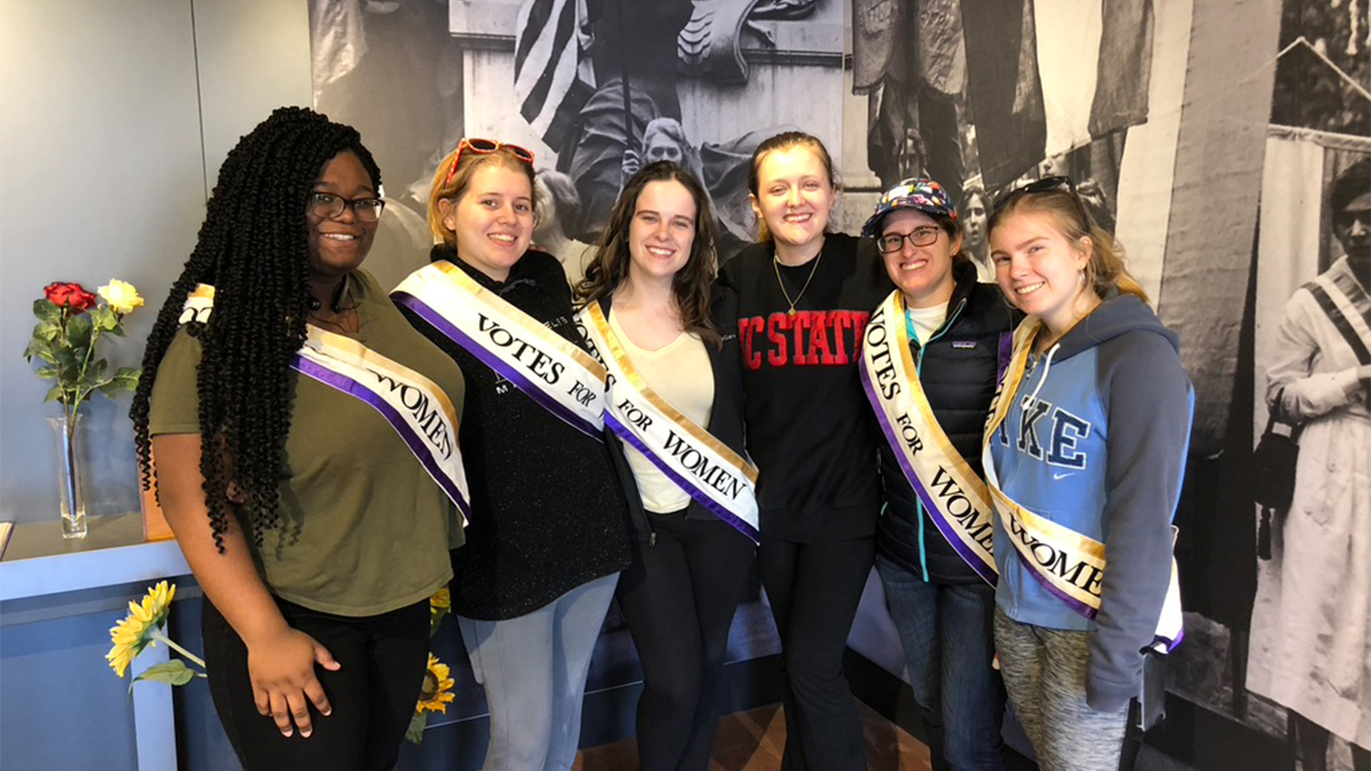 A group of five students from the Women of Welch Village wearing sashes that say "votes for women."
