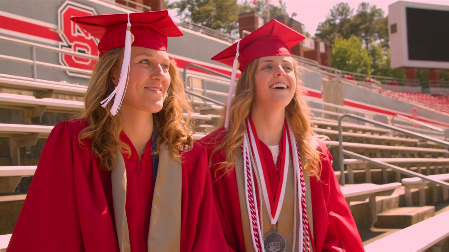 Alyssa McInnis and Emily Southard in their red graduation robes, sitting in the stands of Carter Finley Stadium