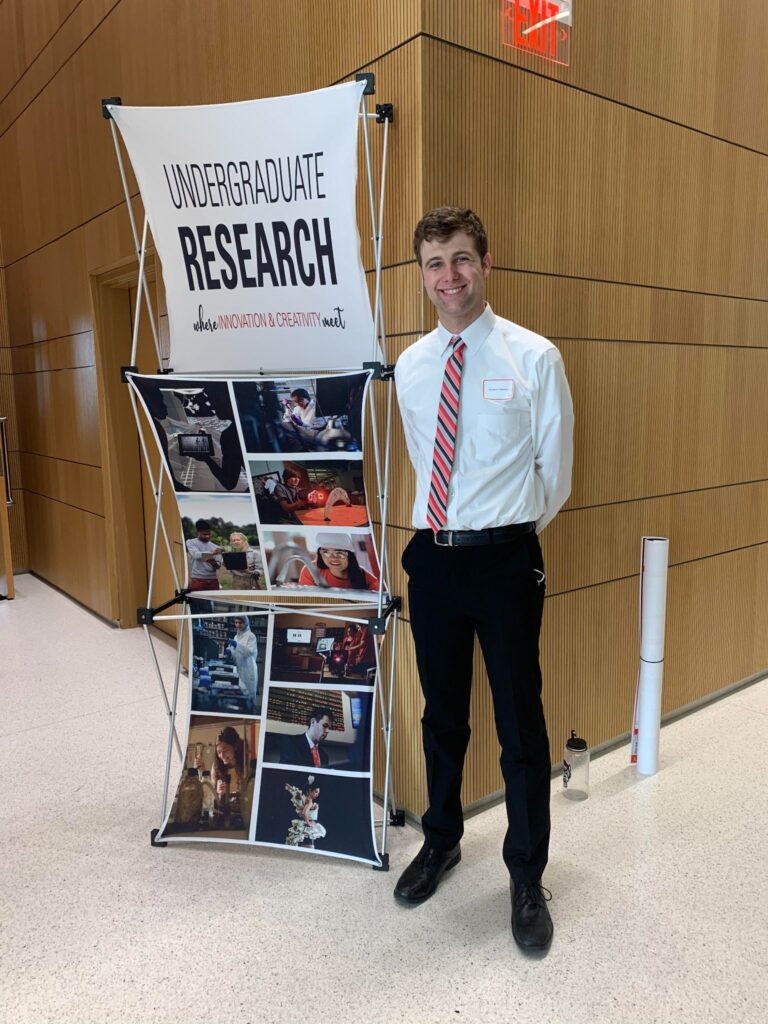 Ben Masters in front of a standing sign displaying the words "Undergraduate Research" with a collage of photos