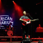 BJ Barnham plays guitar on stage in front of the lit up sign that says "American Aquarium."