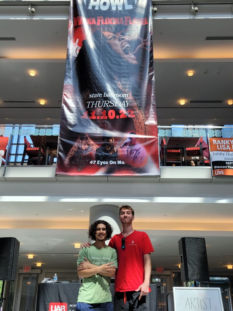 Two students in front of a giant banner promoting the PackHowl concert, dangling from the upper floors in the lobby of Talley Student Union