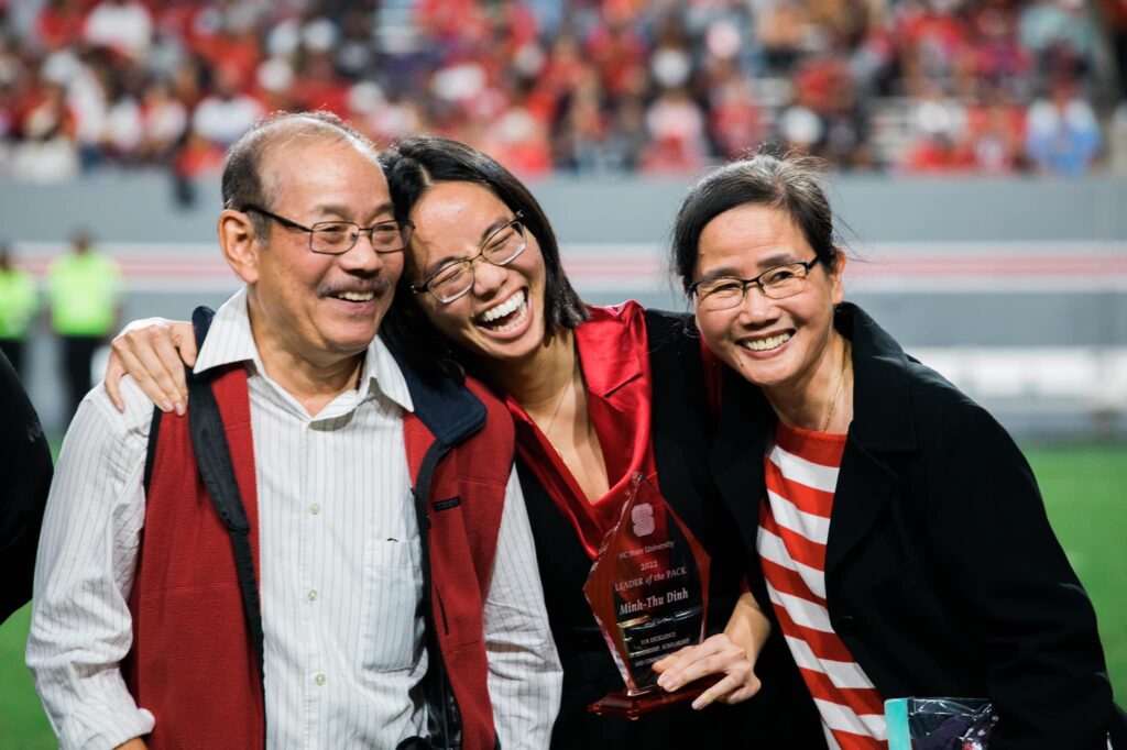 Minh-Thu Dinh and her parents embrace and smile while she holds a plaque and stands on the football field for the Leader of the Pack ceremony.