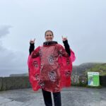 A woman in a red poncho makes wolfie signs with her hands in front of a cloudy scenic overlook