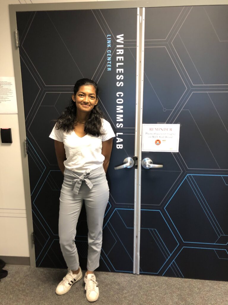 Sucheta Malladi standing in front of blue double doors that read "Wireless Comms Lab Link Center."