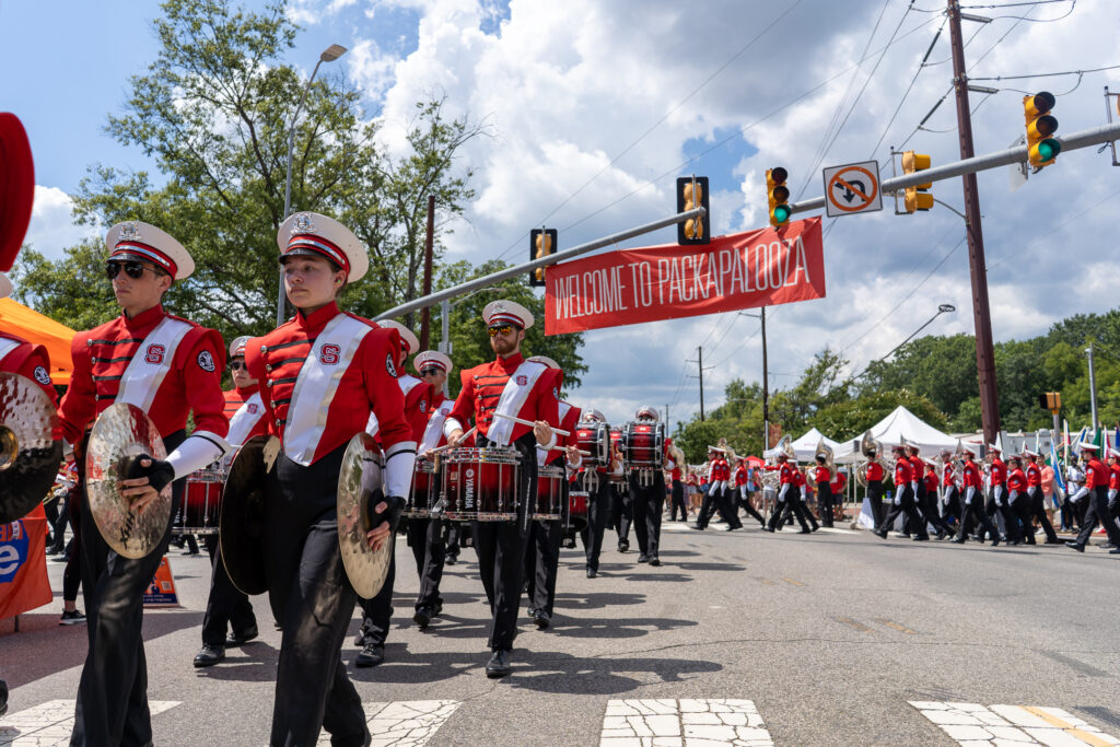 The NC State marching band marches in front of a large banner down Hillsborough Street to kick off Packapalooza 