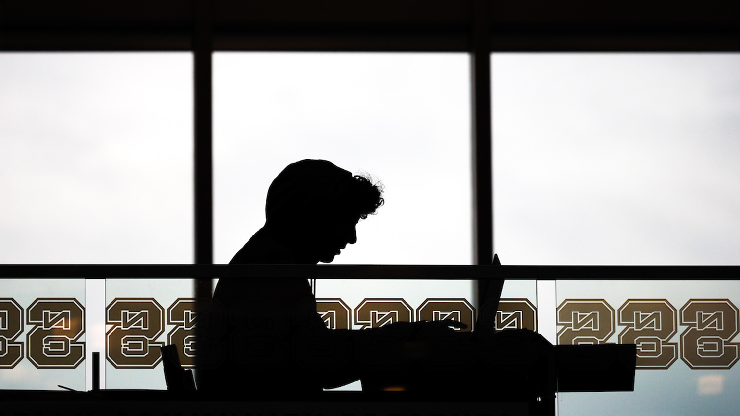 Silhouette of a student studying by a large window with NC State logos along the bottom