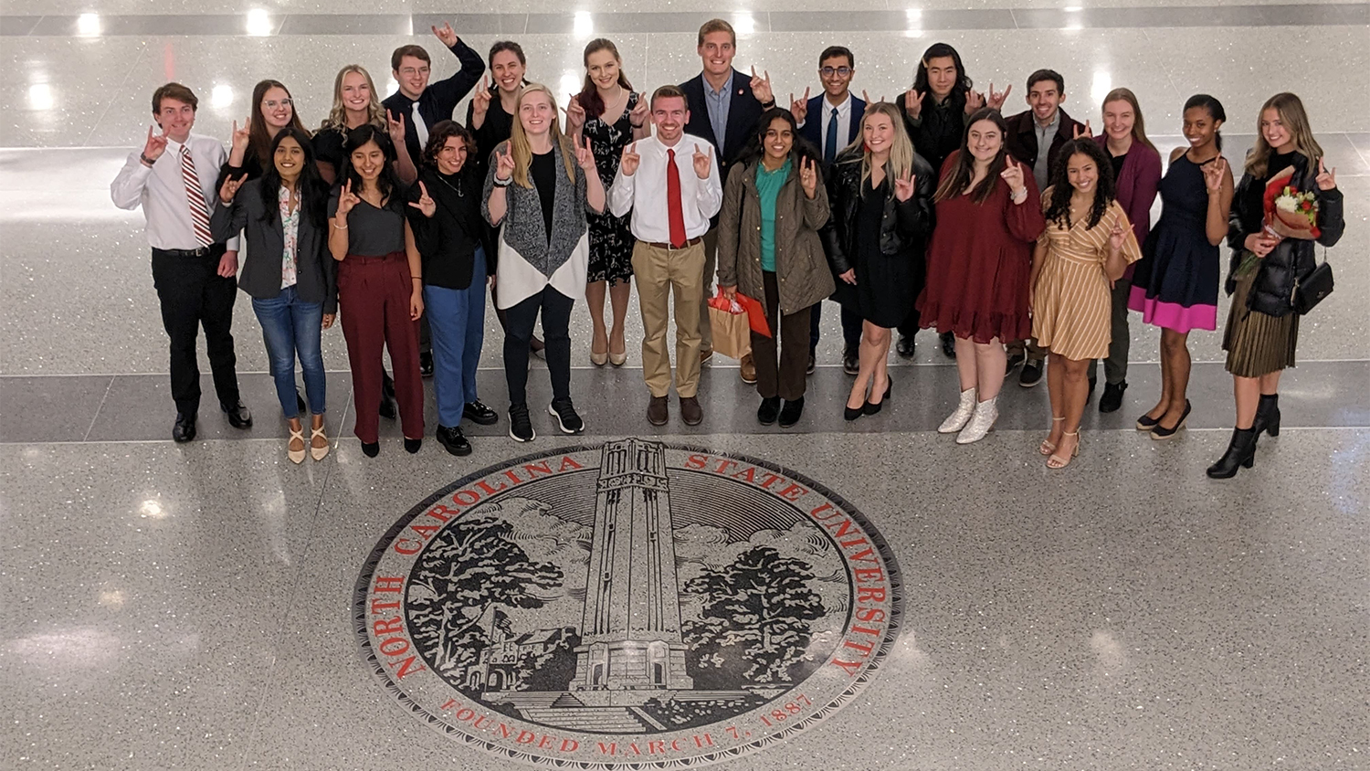 A large group of students stands in front of the NC State seal on the floor of Talley Student Union