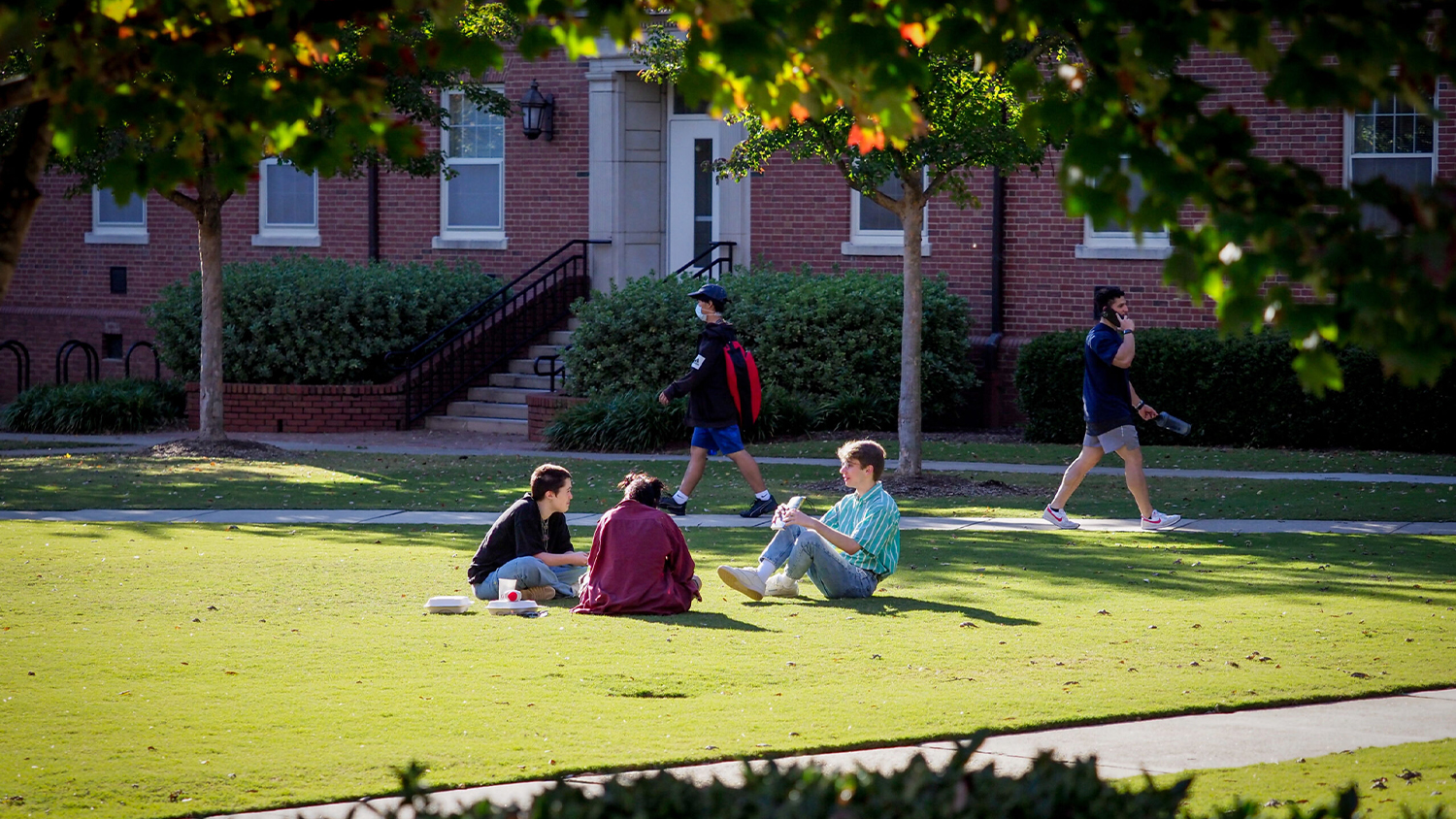 Students sit in a circle on the lawn in front of a residence hall while two other students walk by on the sidewalk behind them