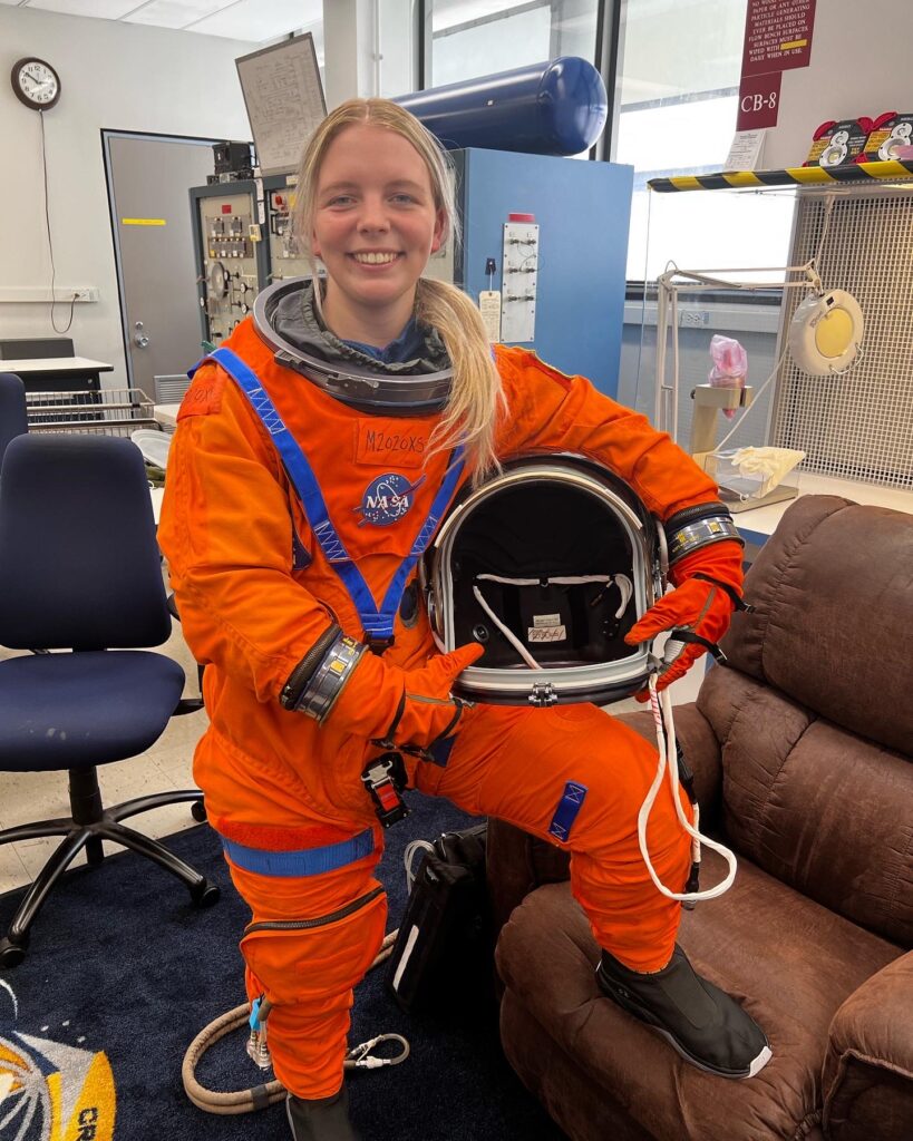 Kierra wearing an orange astronaut uniform and holding a helmet with her foot propped up on a recliner