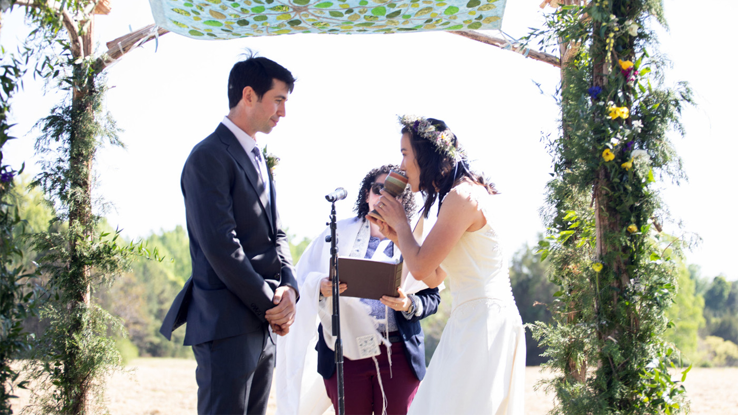 A bride and groom stand in front of a rabbi, while the bride drinks from a cup