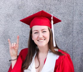 Rylee Young in her red graduation robes making a wolfie sign by the Belltower