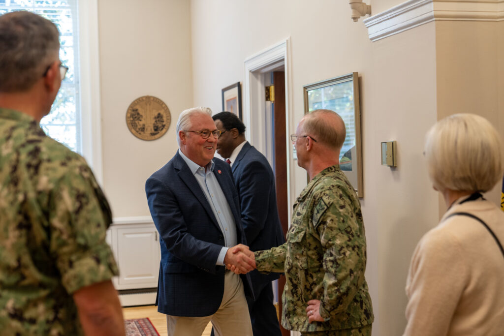 Admiral Caudle shakes hands with Chancellor Randy Woodson in his office, surrounded by other people