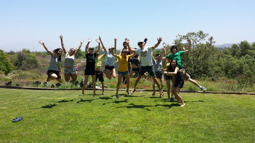 A group of students jump for joy on a grassy field in front of a marsh