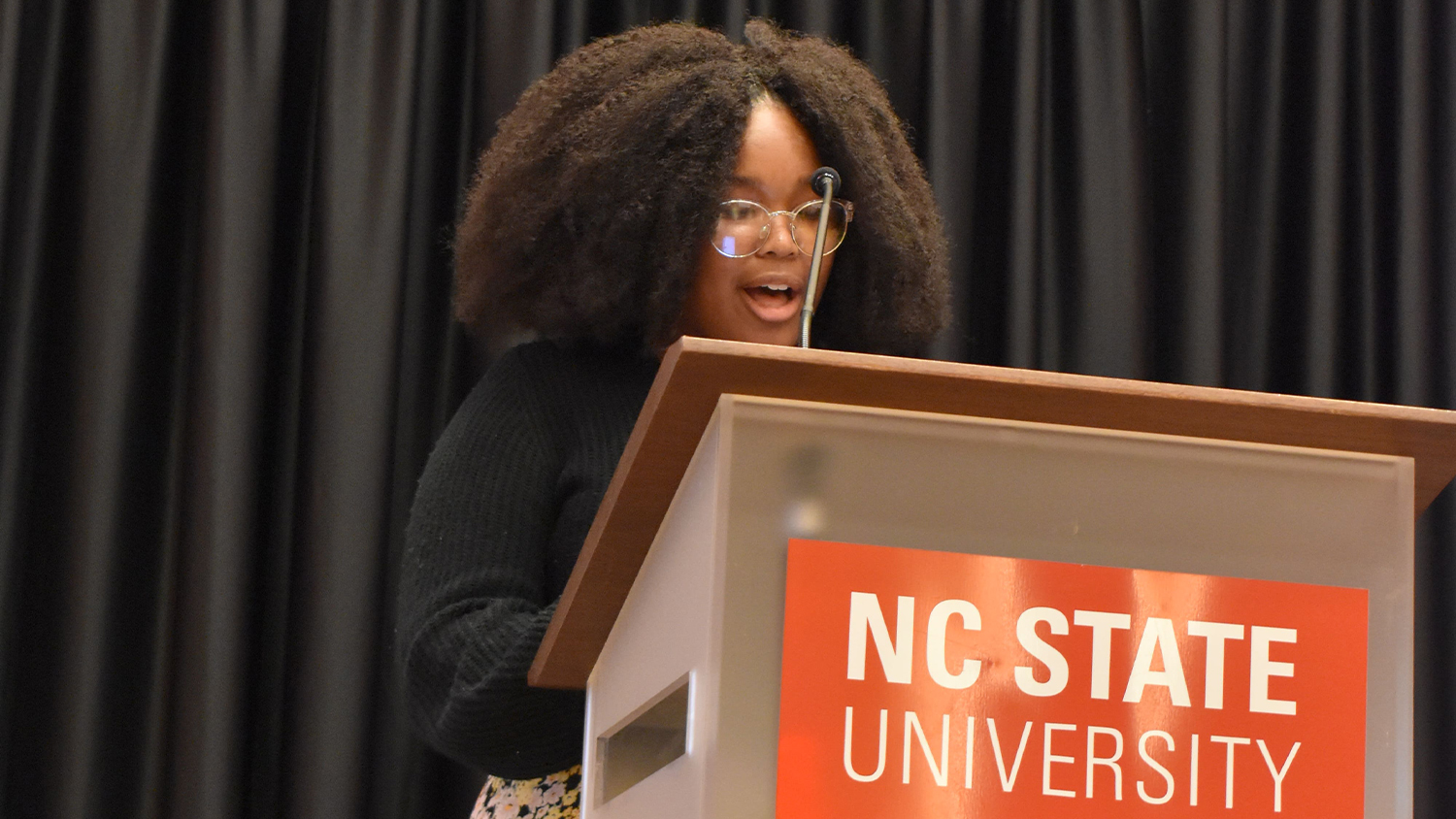 Naudia McKoy speaking at a podium with the NC State logo on it