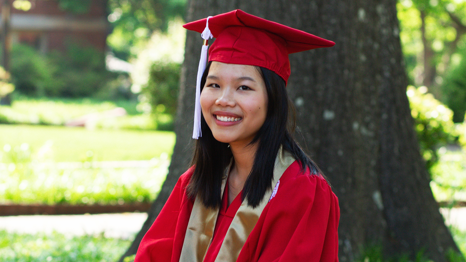 Skye Pham in her red graduation robes in front of a tree on the Court of Carolina
