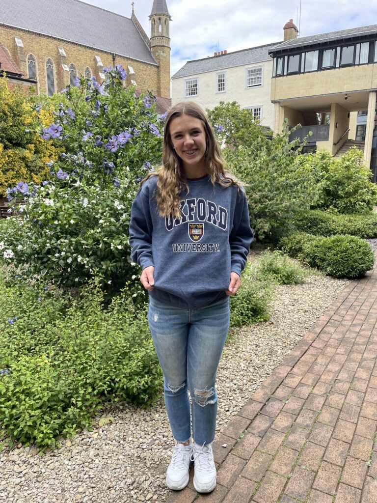 O'Connor standing in a courtyard at Oxford University and wearing a blue sweater with the name Oxford University on it.