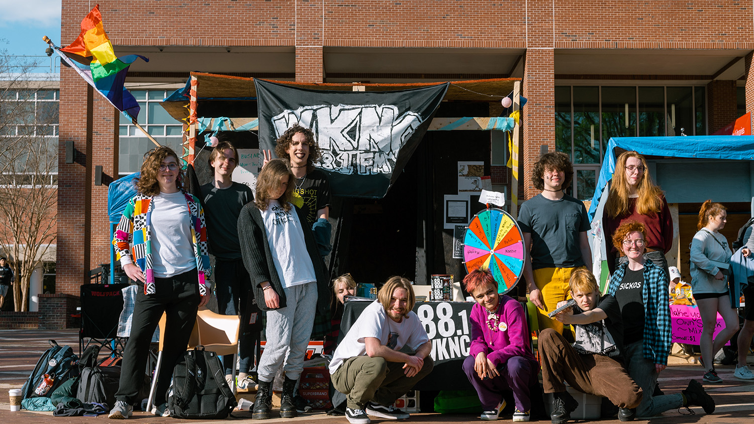 A group of students sit in front of a wooden structure with a black and white flag that reads "WKNC"