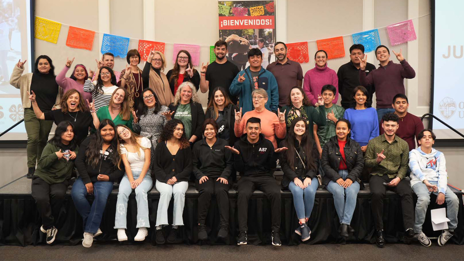 A large group of students and professionals pose for a photo in front of a banner that reads "Bienvenidos!"