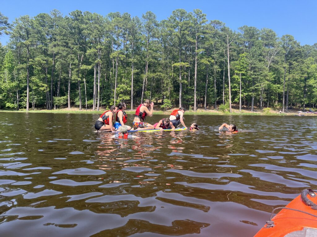 Students work as a team on a paddleboard in a wooded lake.
