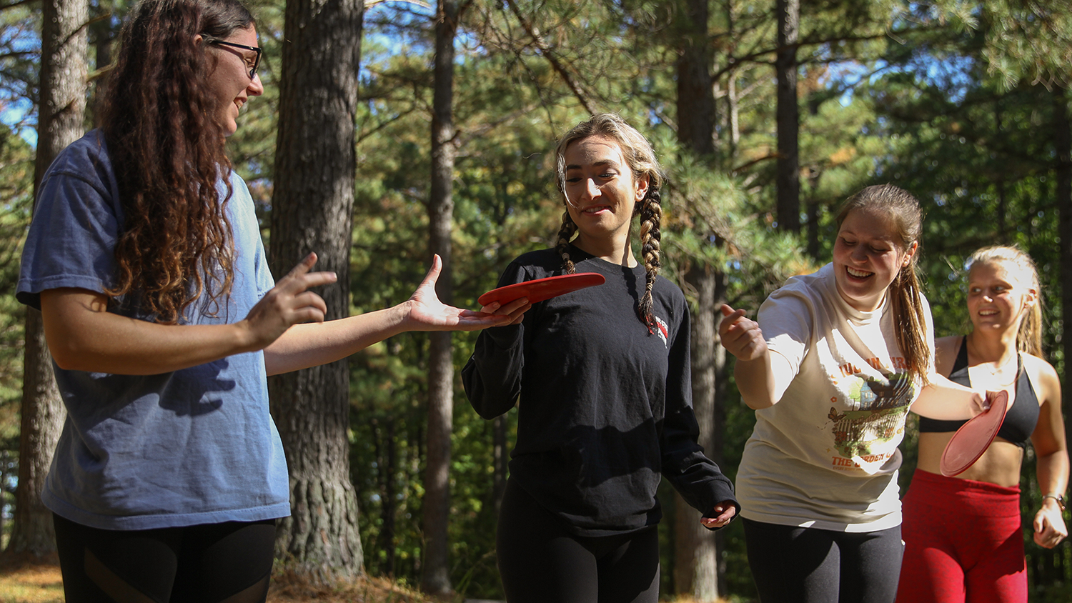 A group of four students in an outdoor environment standing in a line passing discs to each other smiling.