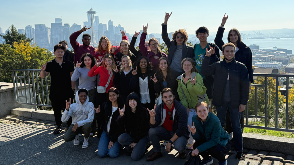 The combined students from the Career Connections: Seattle and Sustainable Cities and Communities: Seattle fall break 2024 trips throw up "Wolfies" in Kent Park, Seattle, WA with the iconic Space Needle set behind them in the city's skyline.