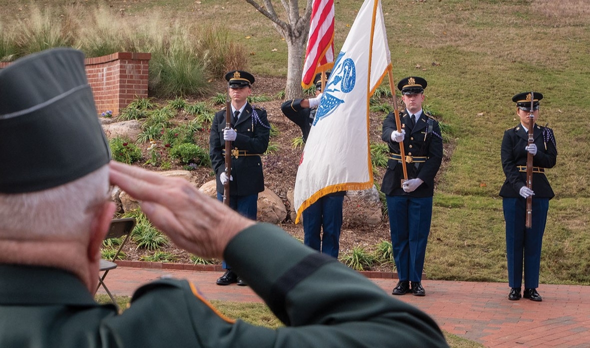 Veteran in military cap saluting in foreground while three-member honor guard in dress uniforms presents American flag and military service flag at NC State Veterans Memorial Garden dedication ceremony.